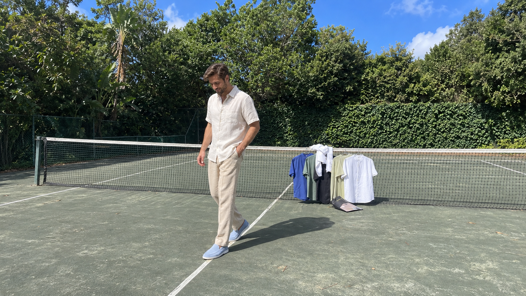 A man in a light short-sleeve shirt, beige trousers and blue slip-on shoes stands on a tennis court with shirts hanging on the net behind him.