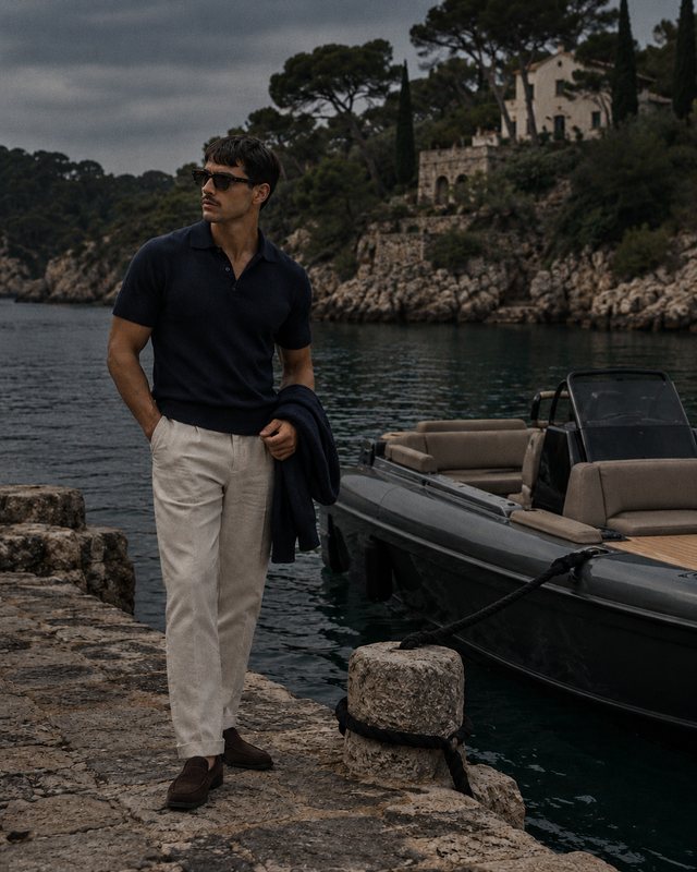 A man in a navy polo and cream trousers stands on a rocky shore beside a dark motorboat.