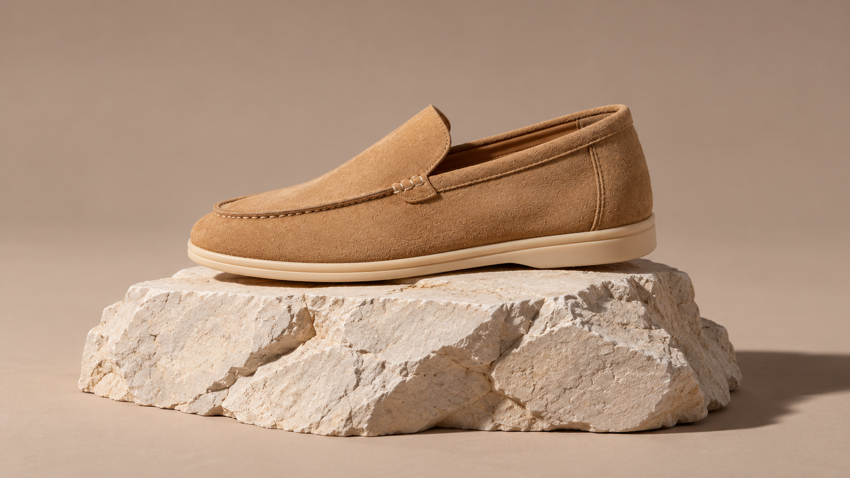 A caramel-coloured loafer with a beige sole is displayed on a pale stone against a warm neutral background.