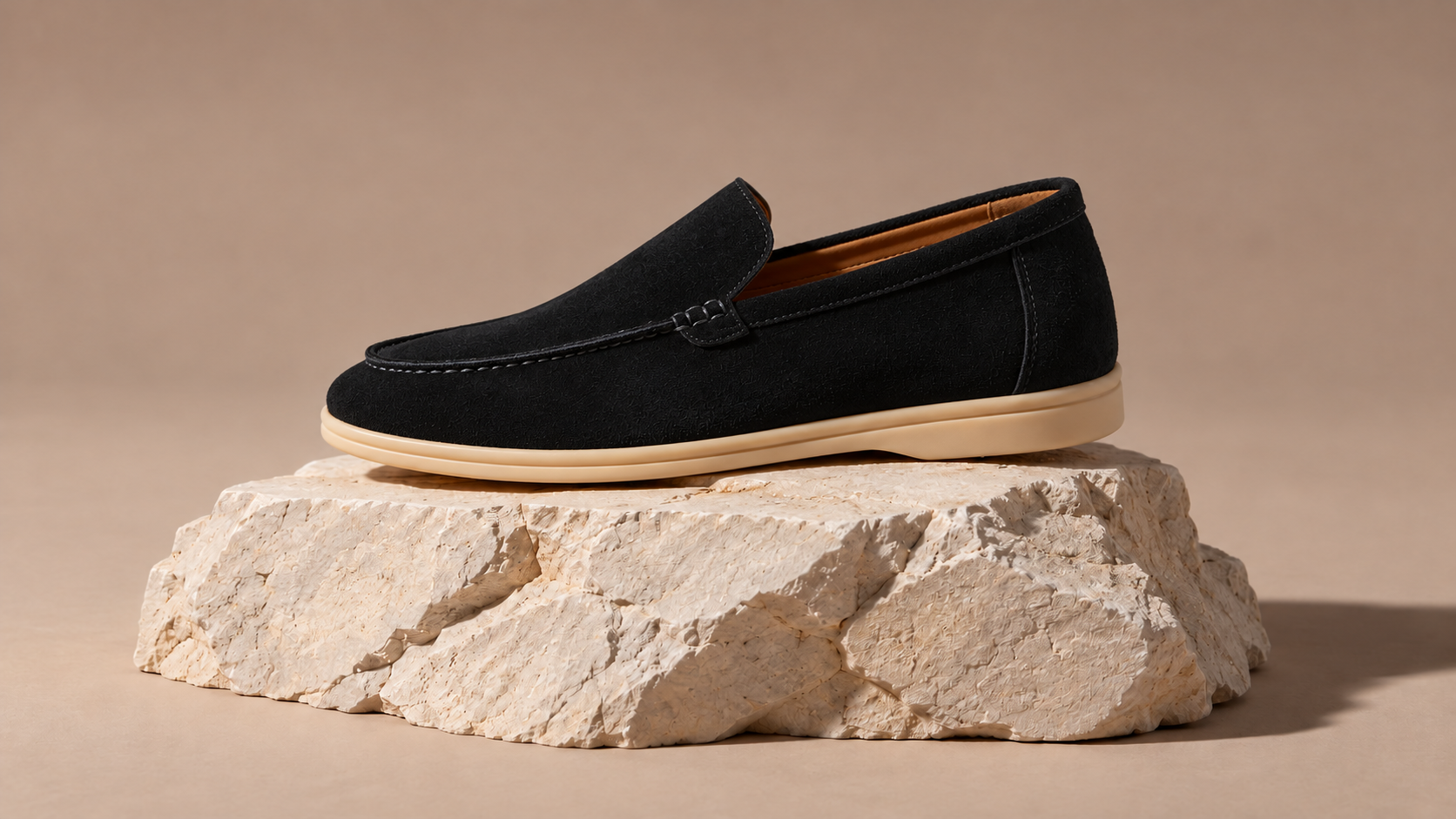 A black suede loafer on a pale stone pedestal.